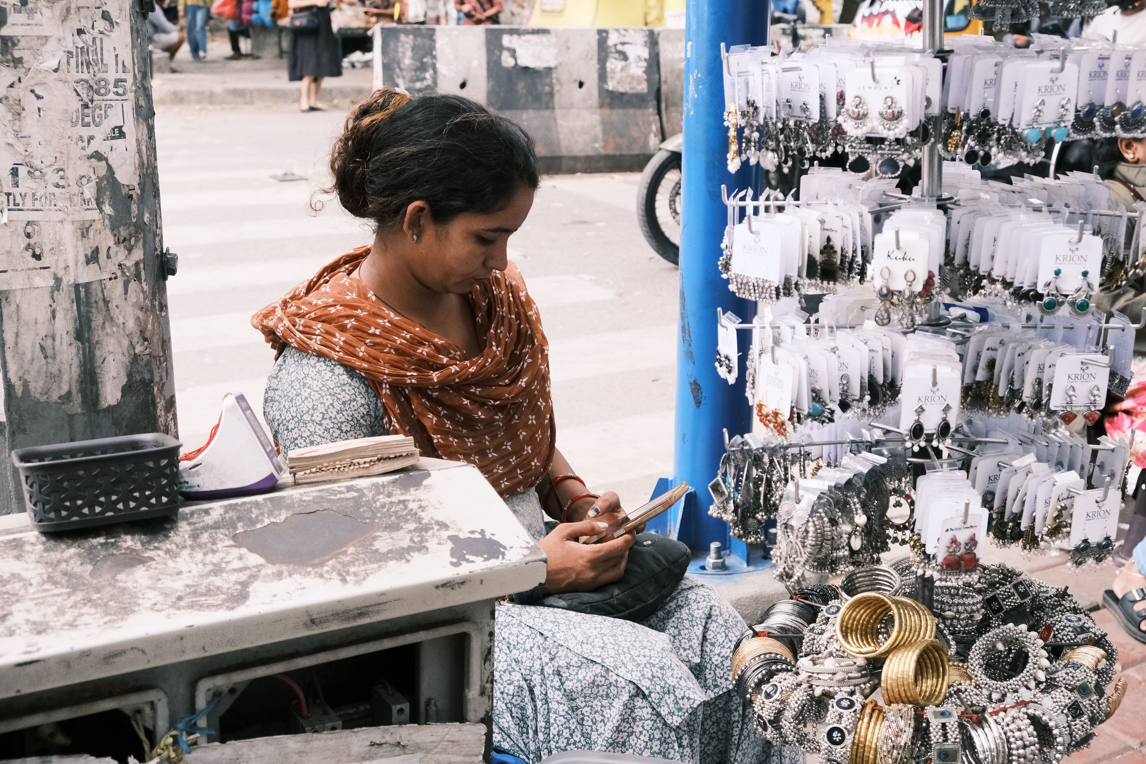 A woman street vendor checking her smartphone at her jewellery stall, hung with earrings and bangles, in a Bengaluru market